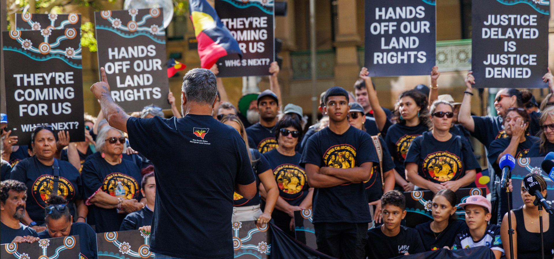 A speaker with back to camera addresses a crowd holding protest signs: 'THEY'RE COMING FOR US', 'HANDS OFF OUR LAND RIGHTS', and 'JUSTICE DELAYED IS JUSTICE DENIED'.