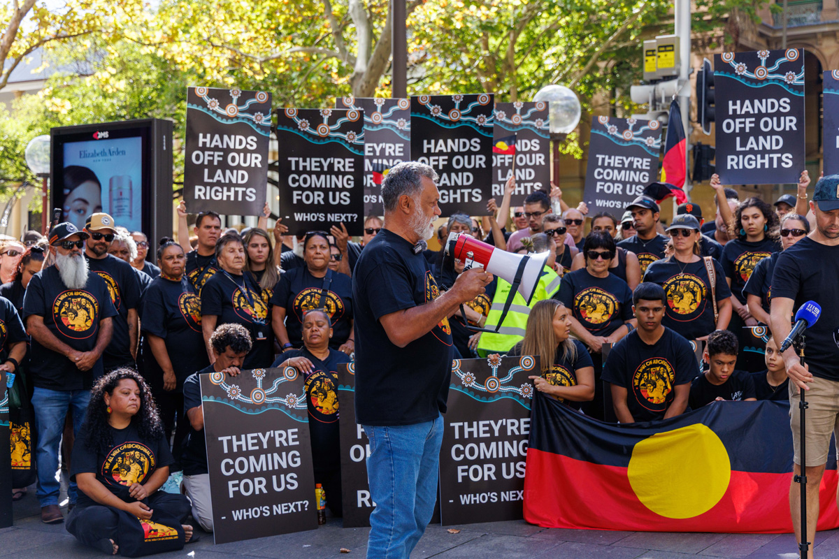 Protest scene with people holding signs saying 'Hands Off Our Land Rights' and 'They're Coming for Us'; a man with a megaphone speaks, Aboriginal flag in foreground.