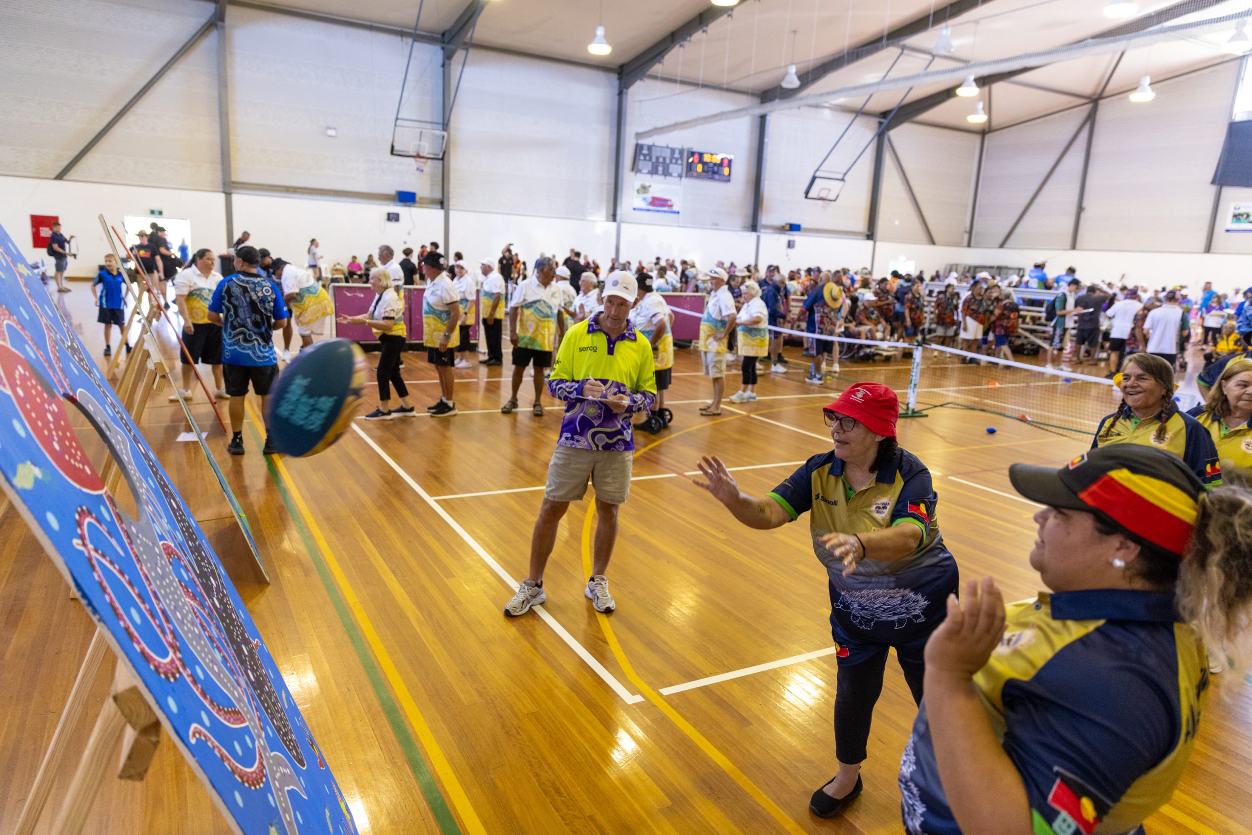 Busy indoor gymnasium filled with people in colorful uniforms, standing in lines and watching a game near a large blue game board on the left.
