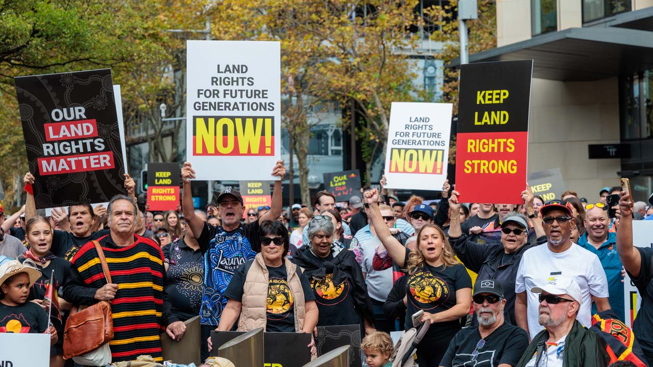 Crowd of protesters holding signs reading 'Our Land Rights Matter' and 'Land Rights for Future Generations NOW!' at an outdoor rally