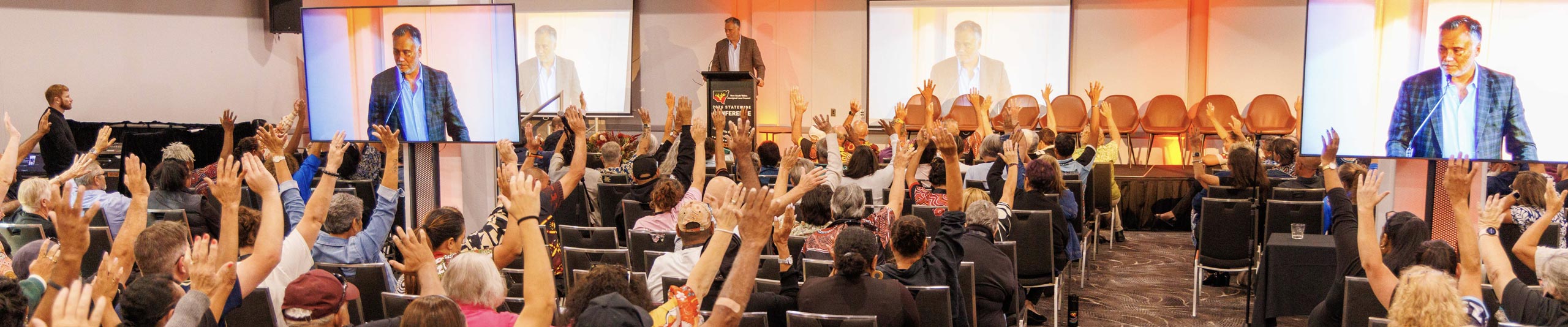 Speaker at podium addressing a large audience in a conference hall as many people raise their hands; screens show the speaker on stage.